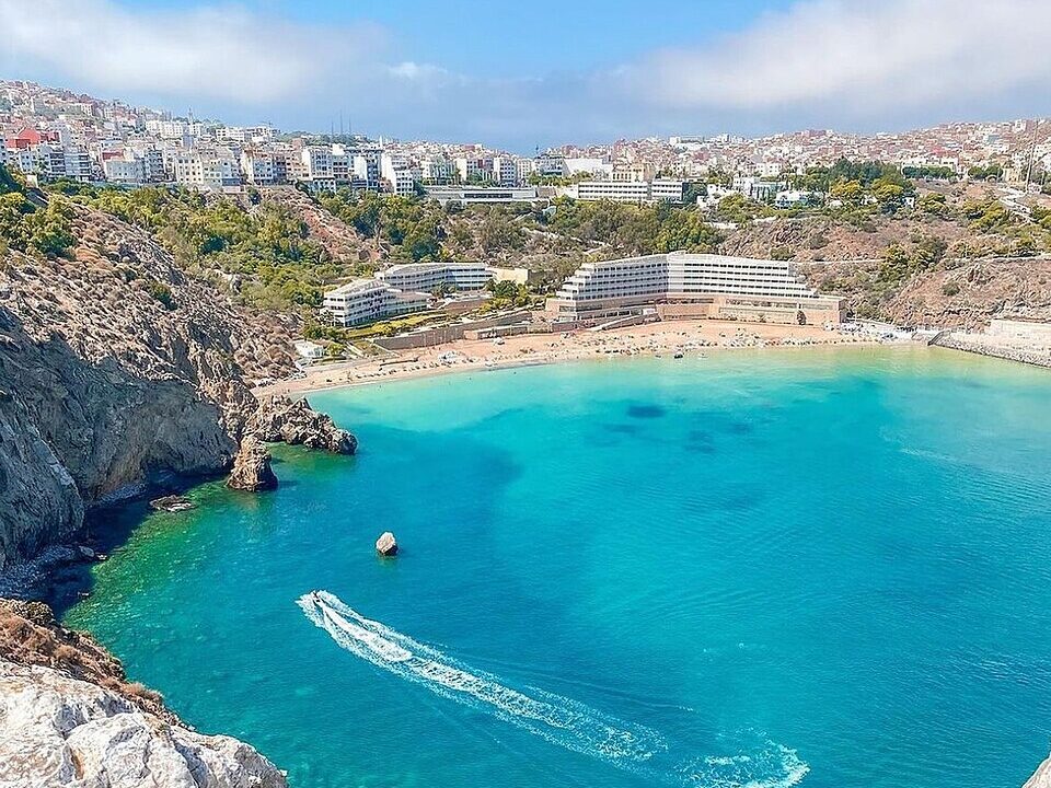 Turquoise water and sandy Quemado Beach beneath Al Hoceima, surrounded by rocky headlands and hillside buildings.